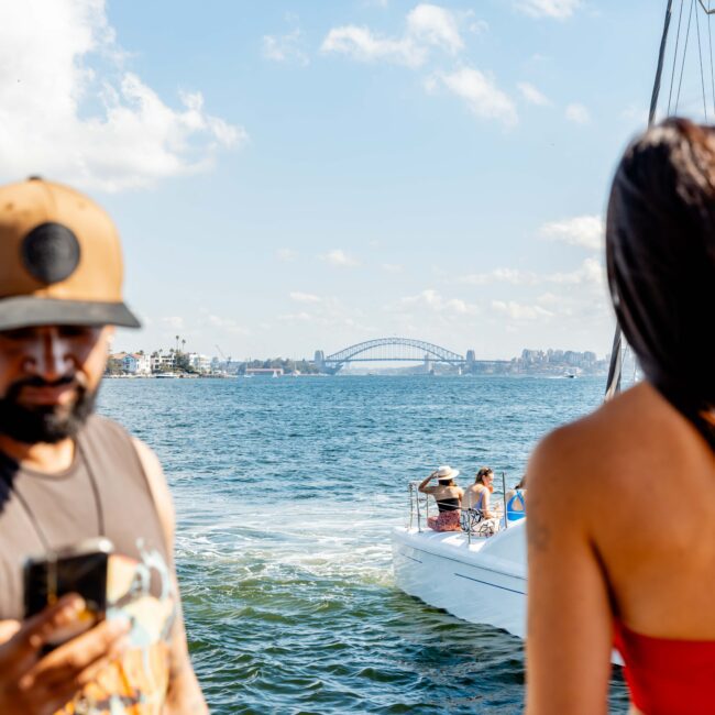 A man looking at his smartphone is in the foreground, and a woman turned away from the camera faces the water. In the background, a boat from The Yacht Social Club is cruising, and a large bridge is visible in the distance.