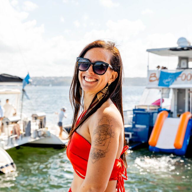 A woman in a red swimsuit and sunglasses smiles towards the camera. She stands on a dock with yachts and people in the background on the water, showcasing The Yacht Social Club Sydney Boat Hire. The sky is clear and sunny, creating a festive, summery atmosphere perfect for boat parties.