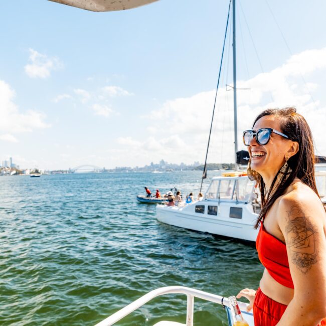 A woman in a red outfit and sunglasses stands on a boat, smiling while looking out over a sunny harbor. Another boat with people aboard is on the water behind her. The scene features a clear blue sky, calm waters, and a distant cityscape, capturing the essence of The Yacht Social Club Sydney Boat Hire.