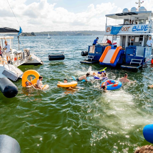 People are enjoying a sunny day out on the water with inflatable tubes and other floatation devices near a luxury yacht. Some are swimming, others are lounging, and there's vibrant action all around. Another boat with people sitting is docked nearby under clear skies.