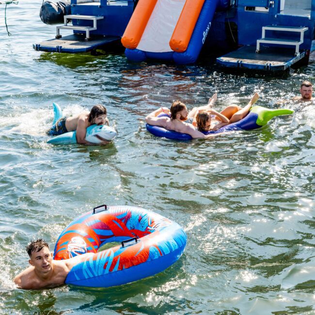 People enjoying a sunny day in the water, swimming and floating on inflatables near a boat. The Yacht Social Club Sydney Boat Hire features a slide set up, and several individuals are using various flotation devices, including an oversized ring and a unicorn float. The background showcases a beautiful lake.