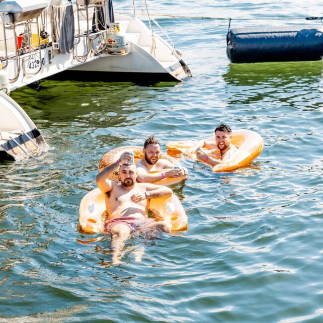 Three men are floating on orange pool floats in a body of water next to a docked boat. The men appear relaxed and are smiling. A buoy is visible in the background. The bright sunny day adds a cheerful atmosphere to the scene. The text "Sydney Harbour Boat Hire The Yacht Social Club" is visible in the bottom right corner.