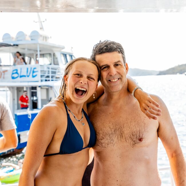 A smiling young woman in a blue bikini and an older man in pink swim trunks stand arm-in-arm on a boat deck, enjoying a Yacht Social Club Sydney Boat Hire. Another boat named "Blue" is visible in the background on the water, enhancing the joyful and sunny atmosphere.