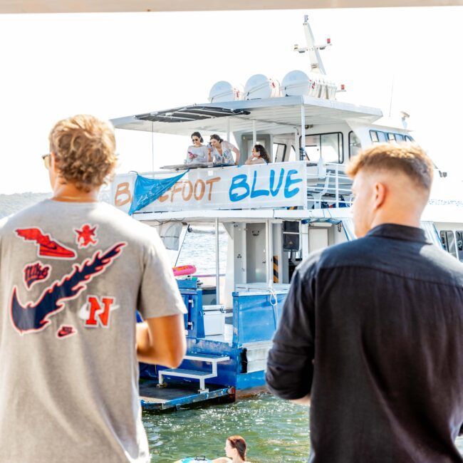 Two people in the foreground stand on a dock, facing a boat named "Barefoot Blue," which is filled with people enjoying the sunny day. The scene depicts a lively and social atmosphere, typical of The Yacht Social Club's boat parties in Sydney, with some on the upper deck and others near the water.