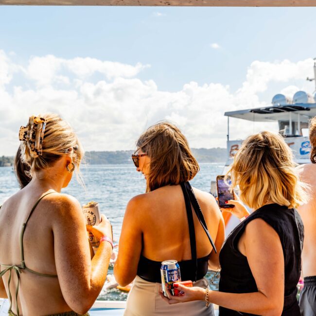 A group of people stands on a boat enjoying a sunny day on the water. They hold cans of drinks and look out at other boats. The Yacht Social Club logo is visible in the bottom right corner, showcasing the exciting atmosphere of Luxury Yacht Rentals Sydney.