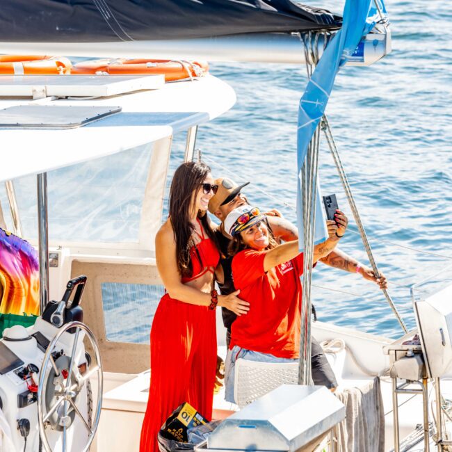 A group of people stands on a sailboat in the ocean. One person in a red dress and sunglasses poses for a selfie with another wearing a red shirt and hat. The boat equipment and blue water are visible in the background, showcasing Luxury Yacht Rentals Sydney by The Yacht Social Club.