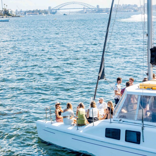 A group of people enjoying a sunny day on the deck of a white catamaran, sailing with The Yacht Social Club Sydney Boat Hire. The boat cruises on a large body of water, with a famous bridge and cityscape visible in the background. The scene appears relaxed and social.