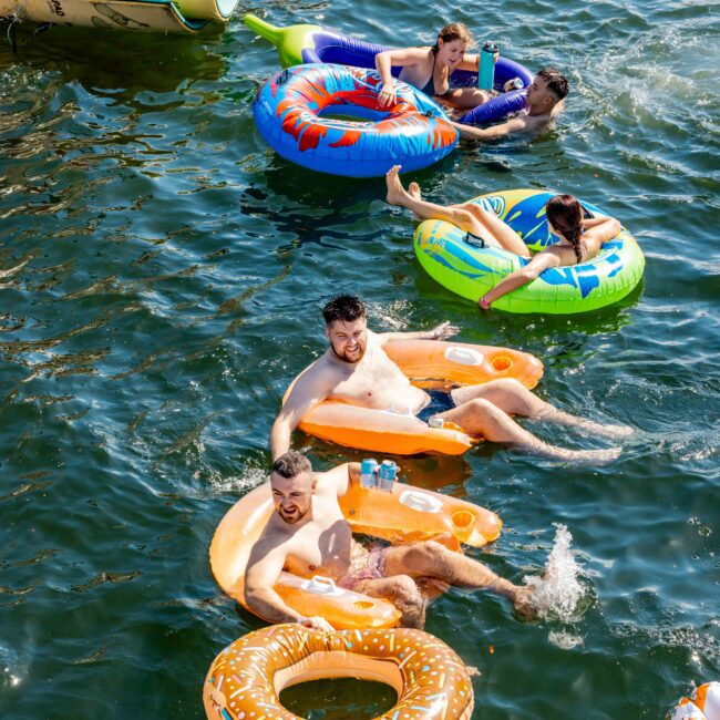 A group of people are floating on colorful, donut-shaped inflatable tubes in the water. Two men in the front are smiling and enjoying drinks, while two others relax behind them. Boats are visible in the background. The Yacht Social Club Event Boat Charters logo is branded on the photo.