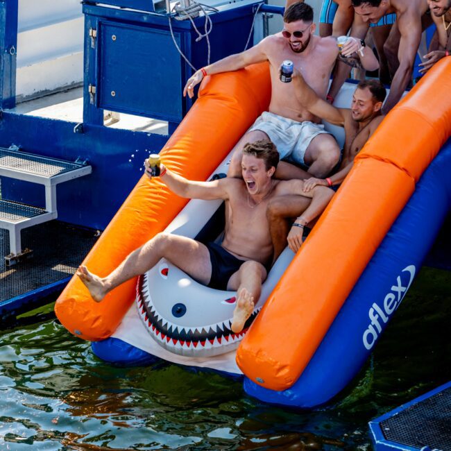 A group of excited men in swim trunks slide down an inflatable water slide attached to a luxury yacht, splashing into the water. One man at the front appears to be shouting joyfully. The event seems festive and fun. The logo "The Yacht Social Club" is visible, making it clear it's part of a Sydney Harbour Boat Hire experience.