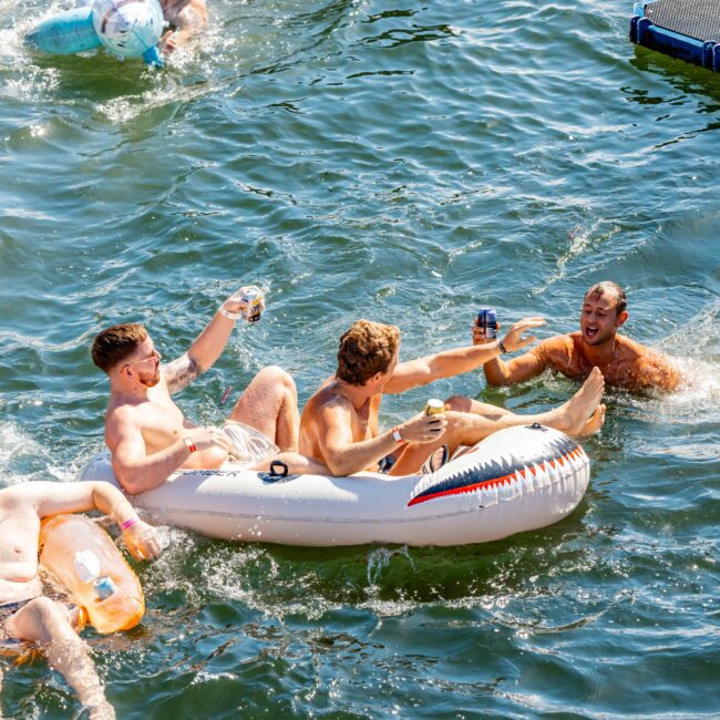 Four men enjoying a sunny day on a lake, three lounging in inflatable devices and one in the water. They laugh, holding drinks, clearly having fun. In the background, more people are on a dock and another inflatable. It feels like a scene from The Yacht Social Club Sydney Boat Hire event.