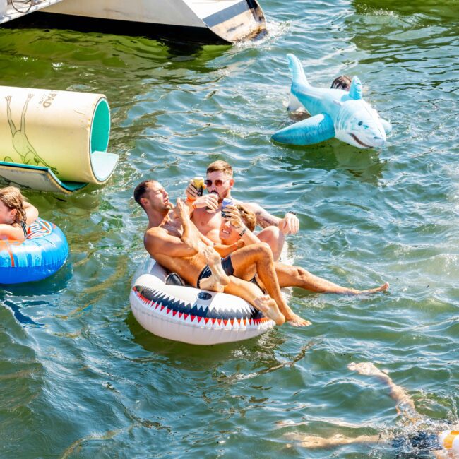 A group of men enjoy a sunny day on the water, lounging on inflatables. One man is seated on a shark-shaped float with others around him. In the background, people can be seen on a boat from Sydney Harbour Boat Hire. Various colorful floats and toys are scattered around in the water.