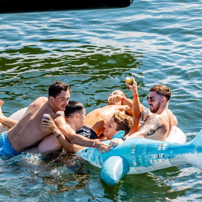 A group of four men are enjoying themselves in a body of water near their Luxury Yacht Rentals Sydney. They are playfully clinging to each other and a large inflatable shark float, holding drinks and having fun on a sunny day.