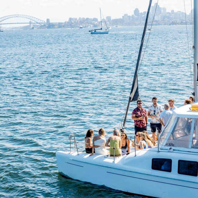 A group of people enjoying a sunny day on a sailboat with a picturesque view of a bridge and city skyline in the background. The boat glides on calm water, with a few other boats in the distance. The logo "The Yacht Social Club Sydney Boat Hire" is visible on the bottom right corner.