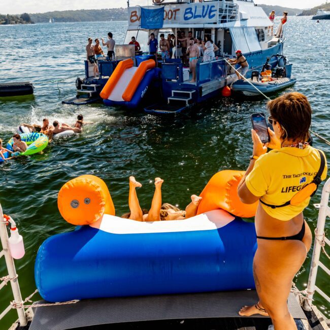 A group of people enjoying a sunny day on a lake, swimming near a boat with slides. One person is taking photos from another boat. Some are lounging on colorful inflatables in the water. Trees and a distant shoreline are visible, evoking the charm of The Yacht Social Club Event Boat Charters in Sydney Harbour.