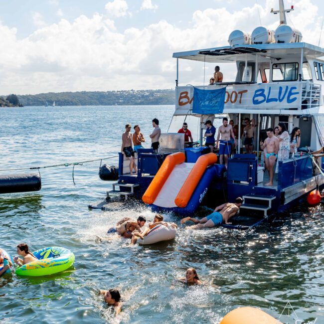 A group enjoys a sunny day on a lake around a party boat named "BLUE," courtesy of Boat Rental and Parties Sydney The Yacht Social Club. Several people swim and use inflatable floats, while others slide into the lake from the boat. The scenic backdrop includes views of the lake and distant shoreline.