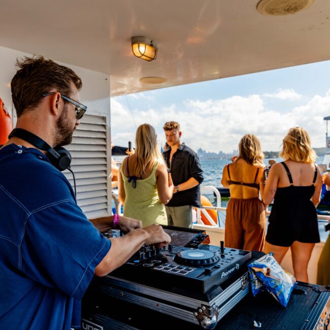 A DJ wearing sunglasses and headphones performs on a boat, using a mixing console. In the background, several people socialize, drink, and enjoy the sunny weather. A lifebuoy is visible on the wall, with "The Yacht Social Club Event Boat Charters" seen at the bottom right corner.