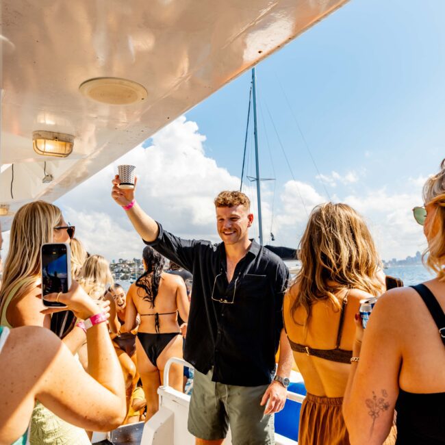 A man in a black shirt and green shorts raises a cup in a toast aboard the "The Yacht Social Club Sydney Boat Hire" yacht, surrounded by women in swimsuits and summer outfits. The yacht is docked, with a blue sky and cityscape visible in the background. "Yacht Social Club" is printed on the bottom corner.