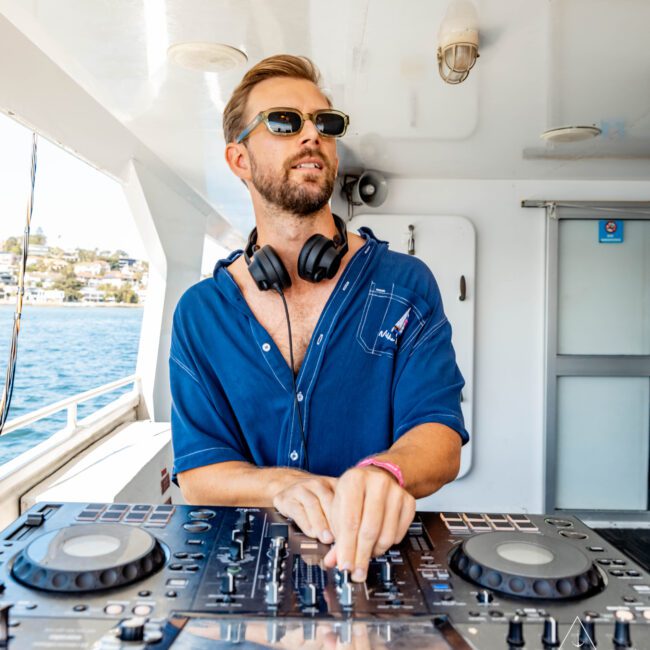 A DJ wearing sunglasses and a blue shirt stands at a mixer console on a luxurious yacht rental in Sydney. With headphones around his neck, he adjusts the controls. The sea and part of a coastal town are visible in the background. The words "Yacht Social Club" are seen in the corner.