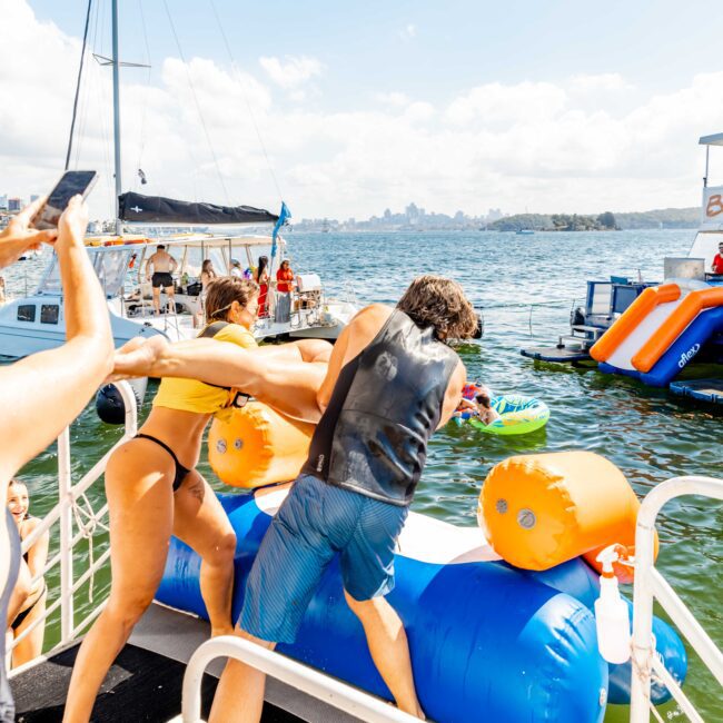 People are enjoying a sunny day on a boat in Sydney Harbour. One person in a yellow bikini and another in blue shorts are playing by pushing an inflatable obstacle while others watch and take photos. In the background, colorful inflatables and the city skyline create a vibrant scene, perfect for Boat Parties Sydney The Yacht Social Club.