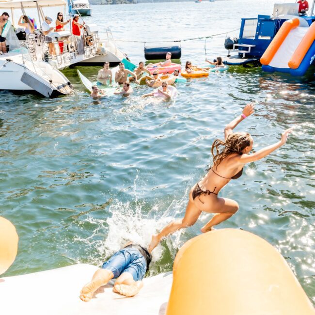 People are enjoying a summer day on the water, with some on boats and others floating in the water. A woman in a bikini is mid-jump off an inflatable slide, while another person splashes into the water below. Inflatable rafts and a slide are visible. The Yacht Social Club Sydney Boat Hire makes it even more fun!