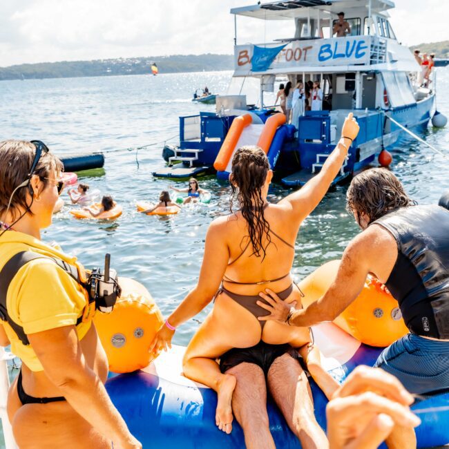 A woman on an inflatable water obstacle gives a thumbs up while a man supports her. Nearby, people play in the water near a boat with "Booze Cruise" on a banner. It's a sunny day, and everyone appears to be having fun, enjoying The Yacht Social Club Event Boat Charters experience.