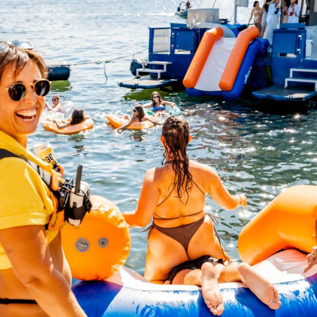 A group of people enjoying a day on the water with inflatables and a large boat from The Yacht Social Club Sydney Boat Hire in the background. One person in a bikini is climbing out of the water on an inflatable, while another smiles at the camera. People are floating on inner tubes and socializing.