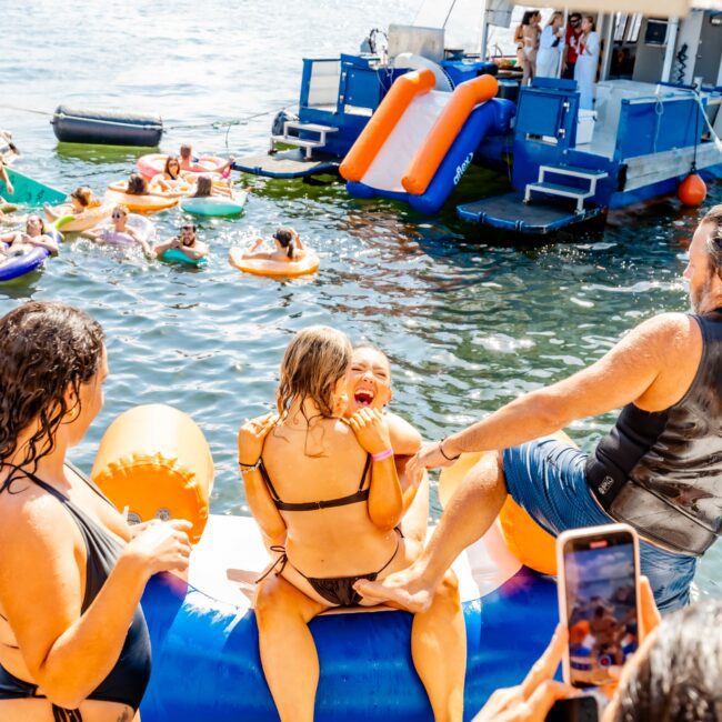 People enjoying a fun day on the water with colorful floaties and inflatable slides. A girl laughs as she sits on an inflatable, while someone captures the moment on their phone. In the background, a boat from The Yacht Social Club Sydney Boat Hire hosts more people participating in water activities.