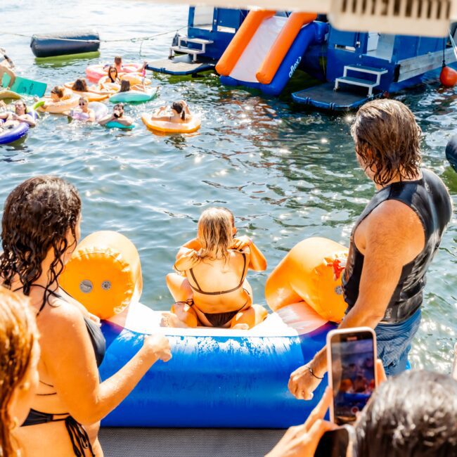 People are enjoying a sunny day on the water with inflatables and a water obstacle course. A person in an orange bikini sits on an inflatable ramp, ready to enter the water, while others watch and take photos. The vibrant and lively scene brings Boat Parties Sydney The Yacht Social Club to mind.