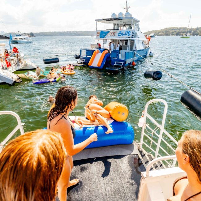 People enjoying a sunny day on a lake with several boats nearby. Two people lie on an inflatable lounger on the deck of one boat. Others are swimming or sitting on the deck, and one boat has inflatable slides. The background features wooded shorelines and other boats from The Yacht Social Club Sydney Boat Hire.