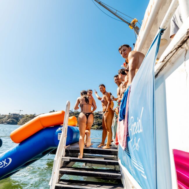 People are standing on the steps of a boat, some in swimwear, with an inflatable slide and water visible in the background. The weather is sunny, and the group appears to be enjoying a day out on Sydney Harbour Boat Hire's luxurious yacht. A banner with the text "Special Effects" is hanging on the boat.