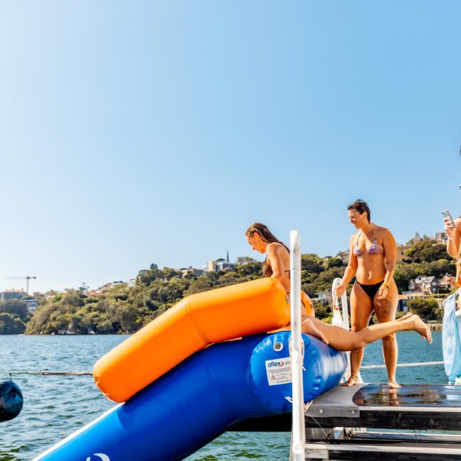 A group of people in swimsuits are on a dock by the water, holding drinks and watching as someone slides down an orange and blue inflatable slide into the lake. In the background, trees and houses dot a hillside under a clear blue sky, giving off The Yacht Social Club Event Boat Charters vibe.