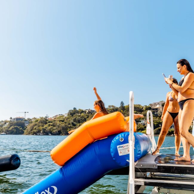 People are enjoying a sunny day on a boat. A woman takes a photo with her phone while another person slides down an inflatable slide into the water. Others stand around, smiling and watching the activity. The backdrop reveals calm water and shoreline, epitomizing The Yacht Social Club Sydney Boat Hire experience.