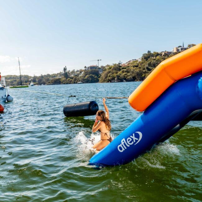 A person splashes into the water from an inflatable slide attached to a boat named "BLUE." Other people are on the boat and in the water, enjoying a sunny day. The background features a hillside with trees and houses, adding to the ambiance of a fun-filled Sydney Harbour Boat Hire The Yacht Social Club event.