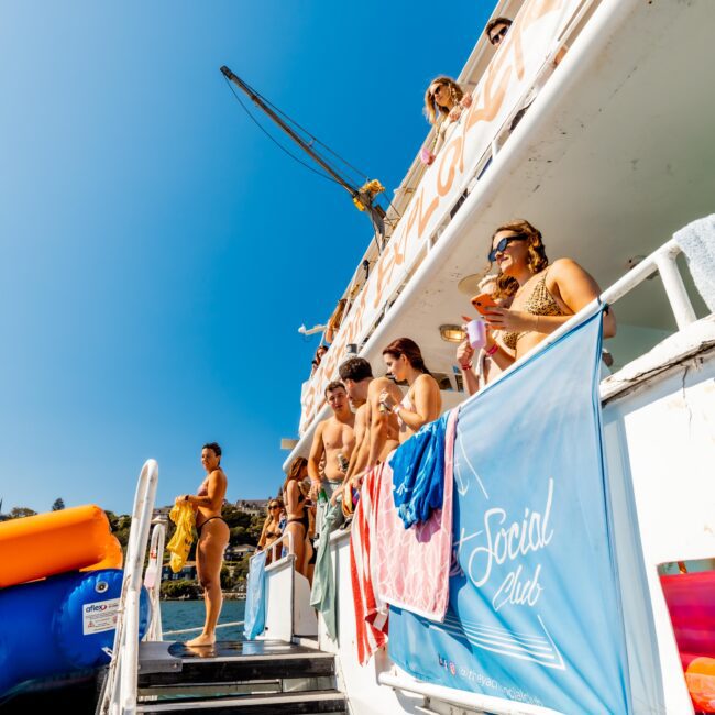 A group in swimsuits is gathered on the deck of a boat, enjoying drinks and sunshine. Next to a blue banner reading "Social Club," an orange slide extends into the water. The clear blue sky adds to the perfect day with Sydney Harbour Boat Hire The Yacht Social Club.