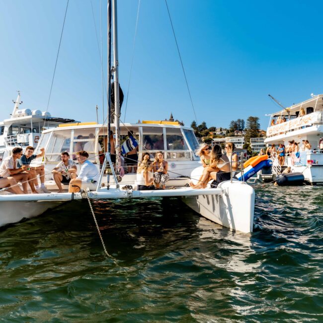 A group of people relaxes on the deck of a white catamaran on sunny, calm waters, surrounded by other boats. Some individuals are seated while others are standing, enjoying the pleasant weather. The background features blue skies, distant trees, and anchored boats—a perfect day with Luxury Yacht Rentals Sydney.