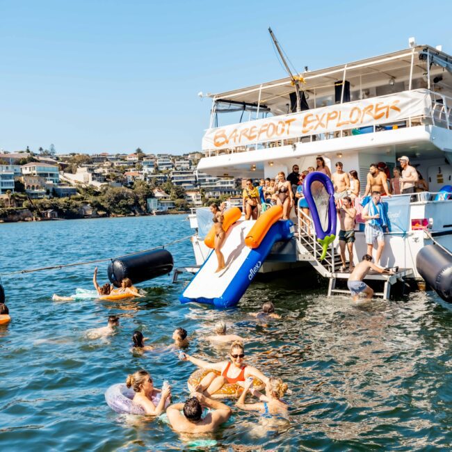 A group of people enjoy a day on the water near a boat named "Barefoot Explorer," courtesy of The Yacht Social Club Sydney Boat Hire. Some are swimming, while others use the boat's slide to enter the water. The boat is anchored, with hillside houses visible in the background.