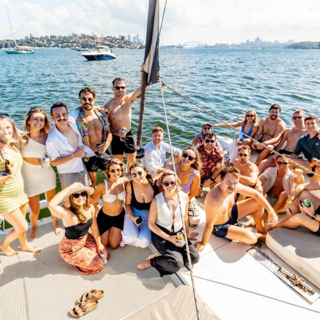 A group of smiling adults enjoys a sunny day on a boat with a scenic city and water backdrop, courtesy of The Yacht Social Club Sydney Boat Hire. Some are sitting while others stand, holding drinks and posing for the photo. The atmosphere is relaxed and festive, with bright, clear skies overhead.