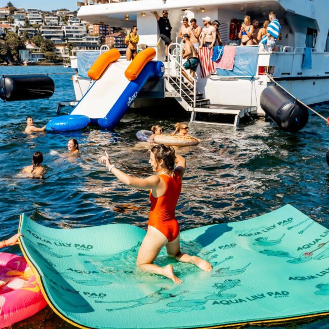 A person in a red swimsuit stands on a floating green pad in the water, posing for a selfie. Nearby, others swim and slide into the water from "Barefoot Explorer," filled with people enjoying the sunny day. The "Yacht Social Club" logo is visible, embodying the vibrant scene of Boat Parties Sydney The Yacht Social Club.