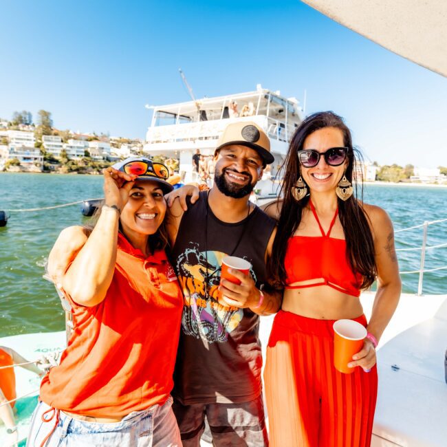 Three friends smiling and enjoying a sunny day on a boat rented from The Yacht Social Club, with houses and another large boat visible in the background. Two women are dressed in red outfits, while the man in the middle is wearing a black hat and T-shirt. They are holding red cups.