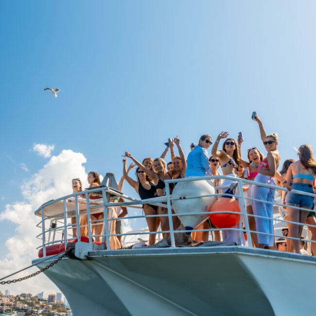 A group of people on the deck of a boat are enjoying a sunny day on the water. Some are taking photos with their phones, while others are raising their arms and smiling. A seagull is flying above them. City buildings are visible in the background, all part of The Yacht Social Club Sydney Boat Hire experience.