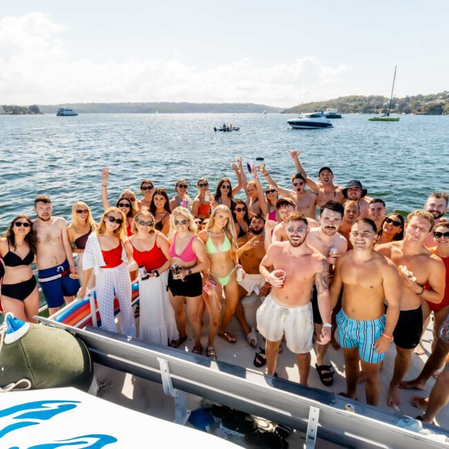 A large group of people in swimwear enjoy a sunny day on a boat. They are smiling, raising their hands, and posing for the camera, surrounded by calm waters and other boats in the distance. The scene conveys a cheerful and celebratory atmosphere, perfectly capturing The Yacht Social Club Sydney Boat Hire experience.