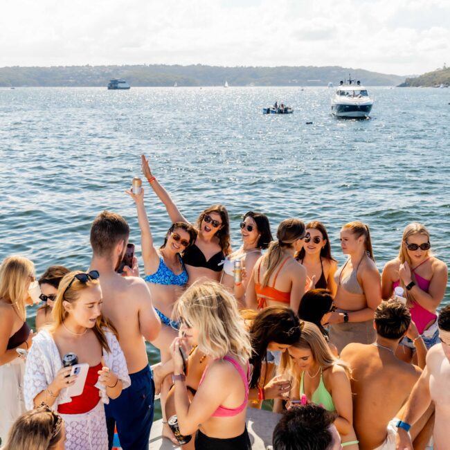 A group of people enjoying a sunny day on a boat. They are wearing swimwear, sunglasses, and summer clothing, holding drinks, and socializing. The boat, part of Luxury Yacht Rentals Sydney, is on calm waters with other boats and land visible in the background.