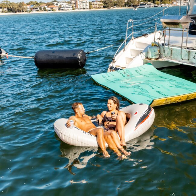 A man and woman relax on an inflatable lounge shaped like a shark in a body of water, with a large boat and paddleboard nearby. They are smiling and enjoying the sunny weather, with buildings and trees in the background. The Yacht Social Club Sydney Boat Hire logo is visible in the corner.