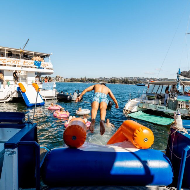 A person in swim trunks dives off a floating platform into a lake, surrounded by colorful floating tubes. Two boats from *The Yacht Social Club* are anchored nearby, adding to the festive atmosphere. The day is sunny and bright, capturing a fun, water-filled gathering.