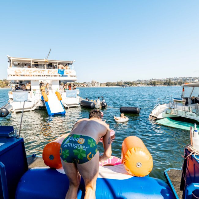 A person wearing brightly colored swim trunks, which read "Budgy Smuggler," leans over an inflatable structure on a boat. Several boats and people enjoying water activities are visible in the background on a sunny day, showcasing the excitement of The Yacht Social Club Sydney Boat Hire.