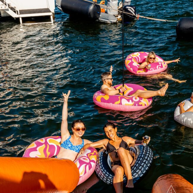 A group of people having fun in colorful inflatable rings on the water, near a docked boat. Two women in the foreground smile and wave, with one holding an alcoholic beverage. The image has a cheerful, summer vibe. The Yacht Social Club logo is visible in the corner, highlighting Boat Parties Sydney.