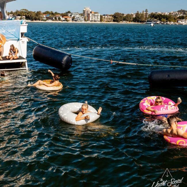 People relaxing on colorful inflatable rings in the water next to a yacht. Two women float on doughnut-shaped rings, while another person relaxes on a plain white ring. The background shows a coastal cityscape under a clear blue sky—it’s the perfect setting for The Yacht Social Club Event Boat Charters.