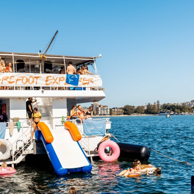 A double-deck boat named "Barefoot Explorer" hosts a lively party with people socializing under a clear blue sky. Two inflatable slides lead into the water, where more guests are swimming and floating on colorful inflatables. A scenic shoreline is visible in the background, synonymous with The Yacht Social Club Sydney Boat Hire.