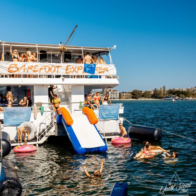 A lively scene on a boat named "Barefoot Explorer" with people enjoying a sunny day. Some are on the deck, others are using a water slide to enter the sea. More people float on inflatable rings nearby. The background shows calm water and a distant shoreline. Perfect for The Yacht Social Club Sydney Boat Hire!