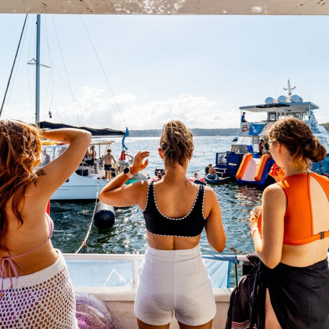 Three women in swimwear stand on a boat deck facing the water and other boats during The Yacht Social Club Event Boat Charters. Water inflatables and people are visible in the background under a clear, sunny sky.